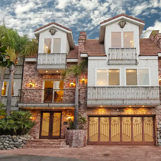 Front view of a two-story modern home with white stucco, brick columns, arched window accents, and decorative wrought-iron gates under a cloudy sky.