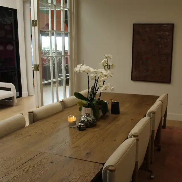A minimalist conference room with a large wooden table, white upholstered chairs, a candle centerpiece, white orchids in a vase, a small sculpture on the tabletop, and tall windows with red frames along one side. A framed artwork hangs on the wall in the background.