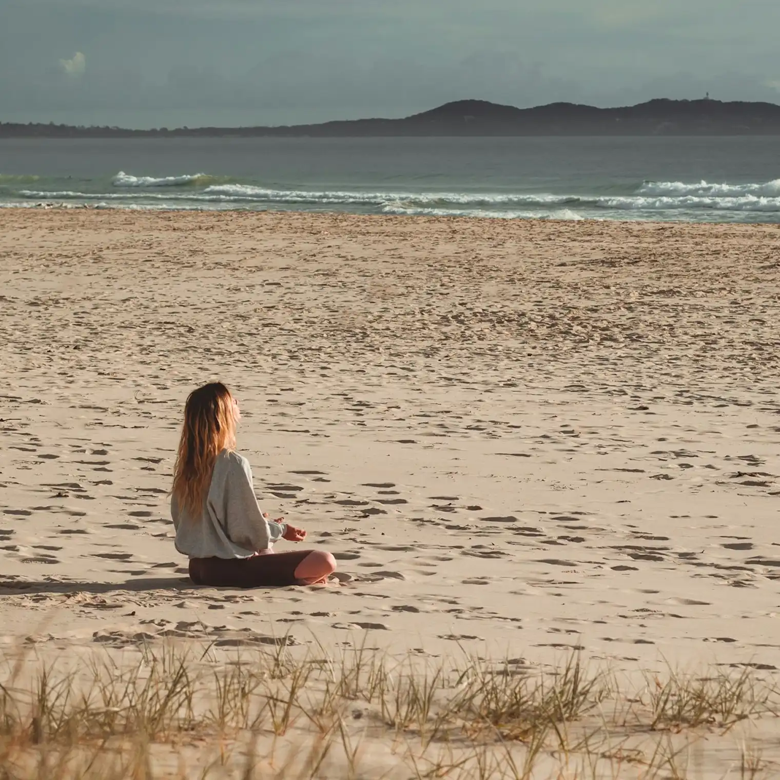 A person sits cross-legged on a sandy beach facing the ocean, with small waves rolling in and dark hills in the distance under an overcast sky.