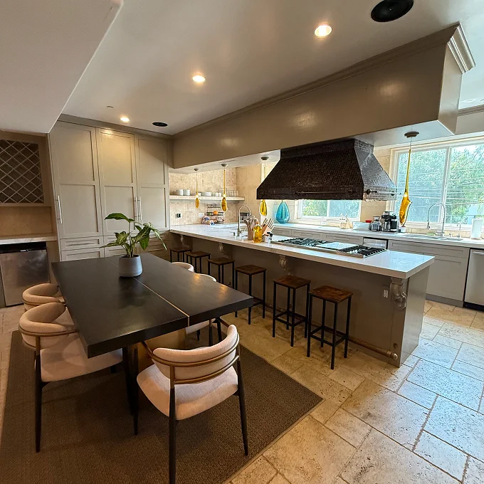 Modern kitchen with a central island, dark bar stools, pendant ceiling lights, and large windows overlooking the yard; white cabinetry and a black range hood are visible.