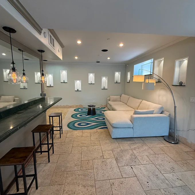 Bright modern living room with beige tile floors, a blue area rug, and a light-colored sectional sofa facing a curved metal fireplace, plus hanging pendant lights and multiple framed wall mirrors.