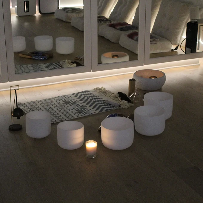 Modern lounge area with a patterned rug, several low white ottomans and stools, a few lit candles, and a large mirrored wall reflecting a seating area with white cushions.