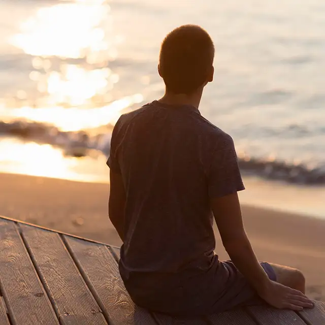 A person sitting on a wooden dock with their back to the camera, silhouetted against a warm sunset over calm water.