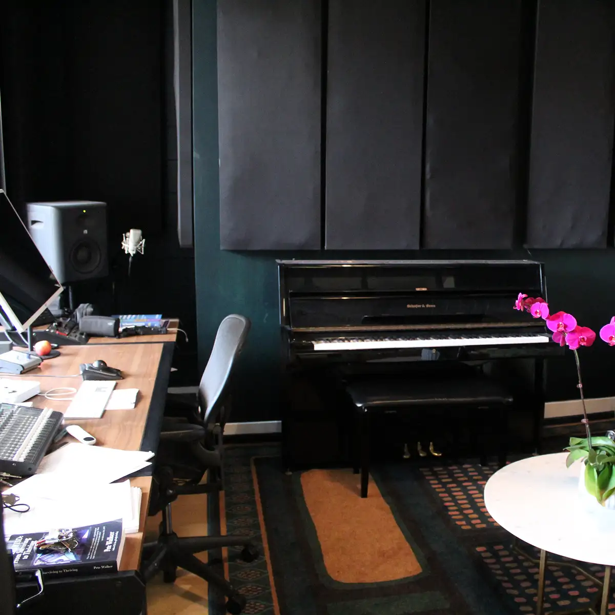 An office studio corner with a desk, computer gear, a black upright piano against the wall, and a bright pink orchid on the table at right.