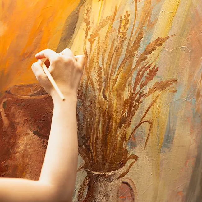 A person’s hand uses a paintbrush to apply warm yellow and orange strokes onto a canvas with textured, tree-like lines.