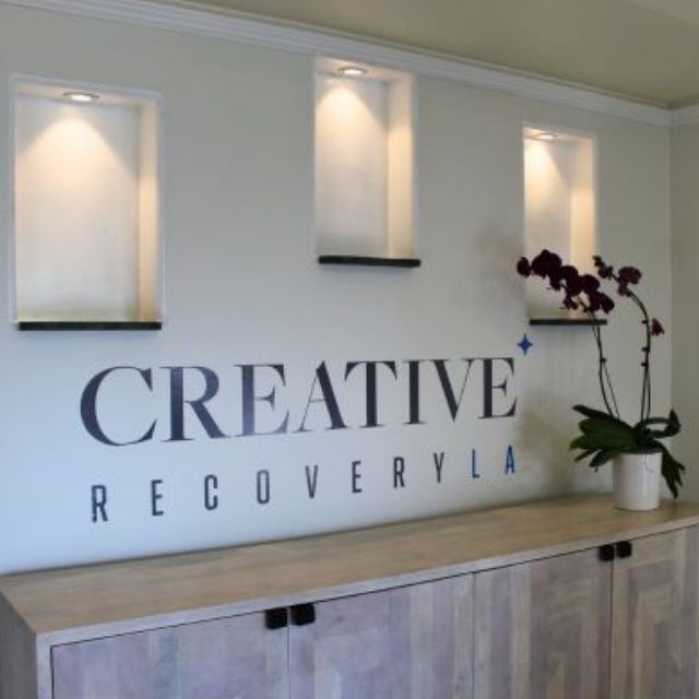 Wall interior with large typography reading “CREATIVE RECOVERYELA” and potted orchids on the right shelf; framed wall lights are mounted above.
