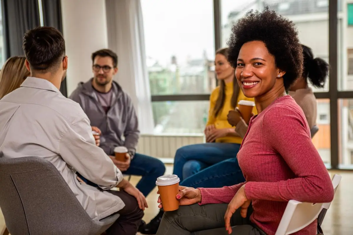 A smiling woman with an afro hairstyle holds a takeaway coffee cup while chatting with colleagues seated in an office lounge; several people in the background hold cups and talk.