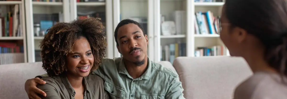 A young couple sits on a couch in a living room, smiling and leaning toward a person in the foreground, with bookshelves in the background.