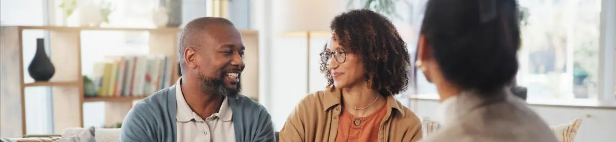 Two people sit and smile while speaking together in a bright, modern room; a third person is visible in the foreground with their back to the camera.