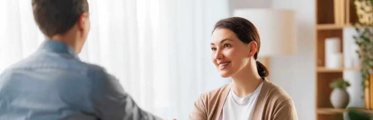 A woman smiles during a conversation while seated indoors, facing a person whose back is visible in the foreground.