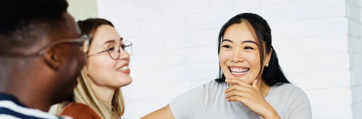 Three people smiling and chatting indoors; one woman in the foreground smiles at the camera with her hand near her chin, with two others partially visible to the left.