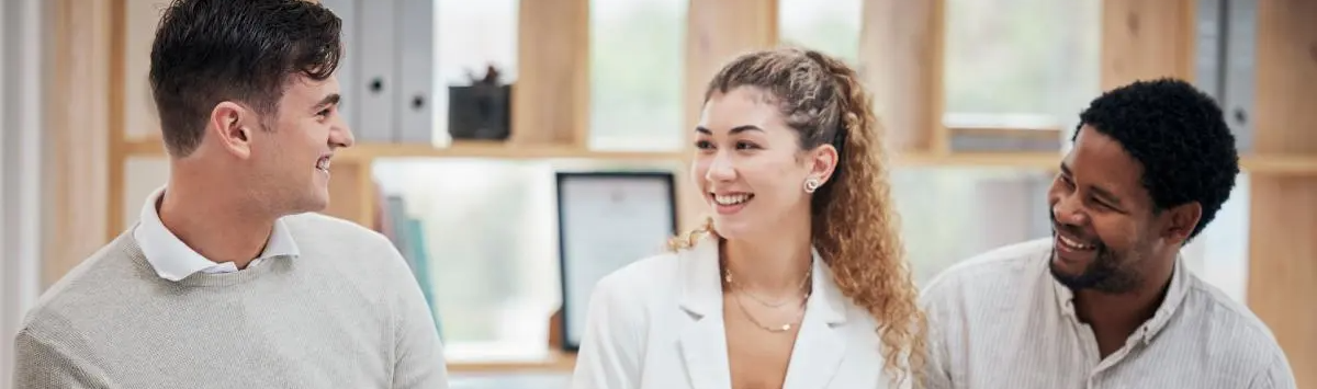 Three coworkers in a bright office smiling and talking together, including a woman in a white blazer and two coworkers in business-casual shirts.