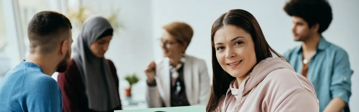A smiling woman in a pink hoodie sits in the foreground while other people talk and work in the background in a bright, indoor room.