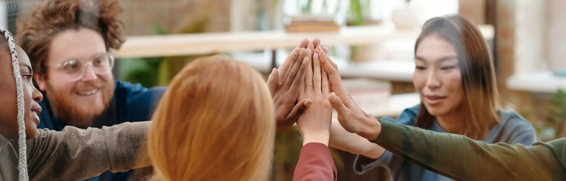 A group of people in a bright living room hold their hands together above a child’s head, smiling and appearing to celebrate a moment of teamwork.