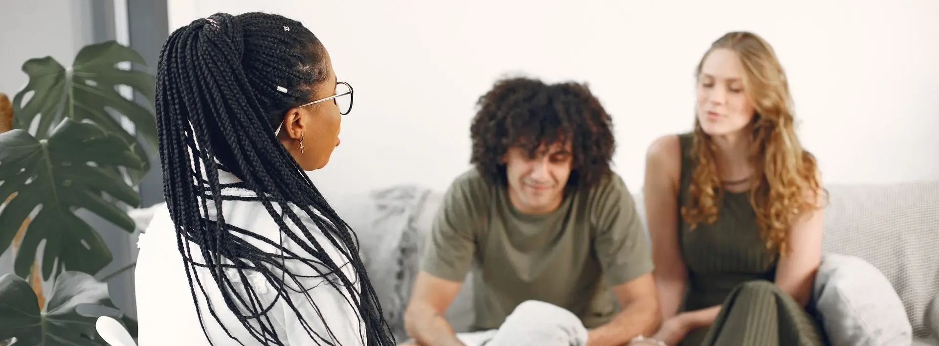 A woman with braided hair and glasses sits and talks while a man and another woman smile in the background on a sofa.