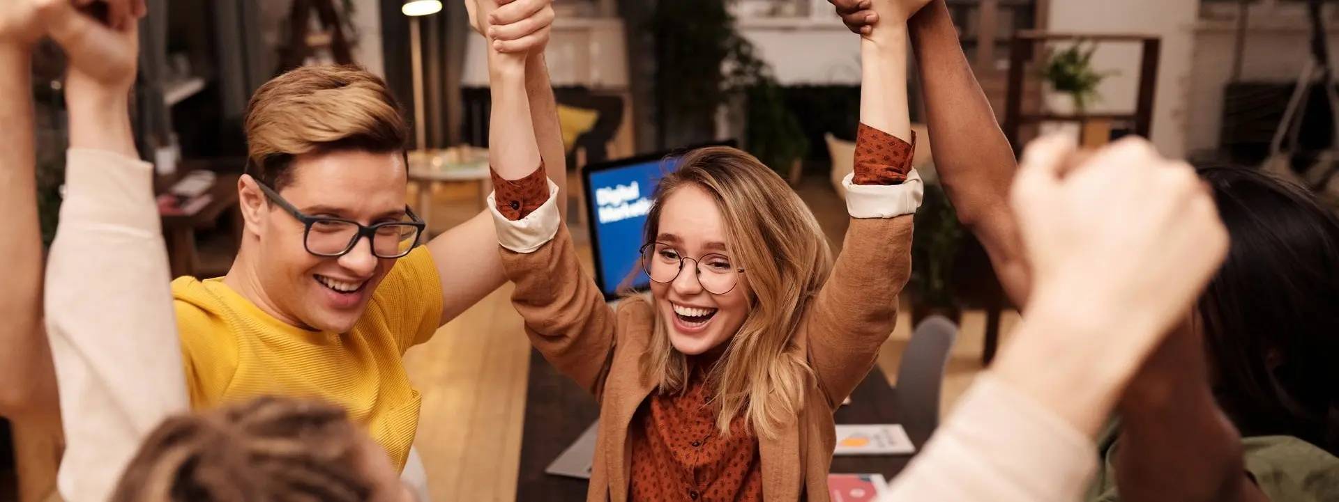 Two young people with glasses in a cozy office or cafe setting shout and raise their arms as others surround them.