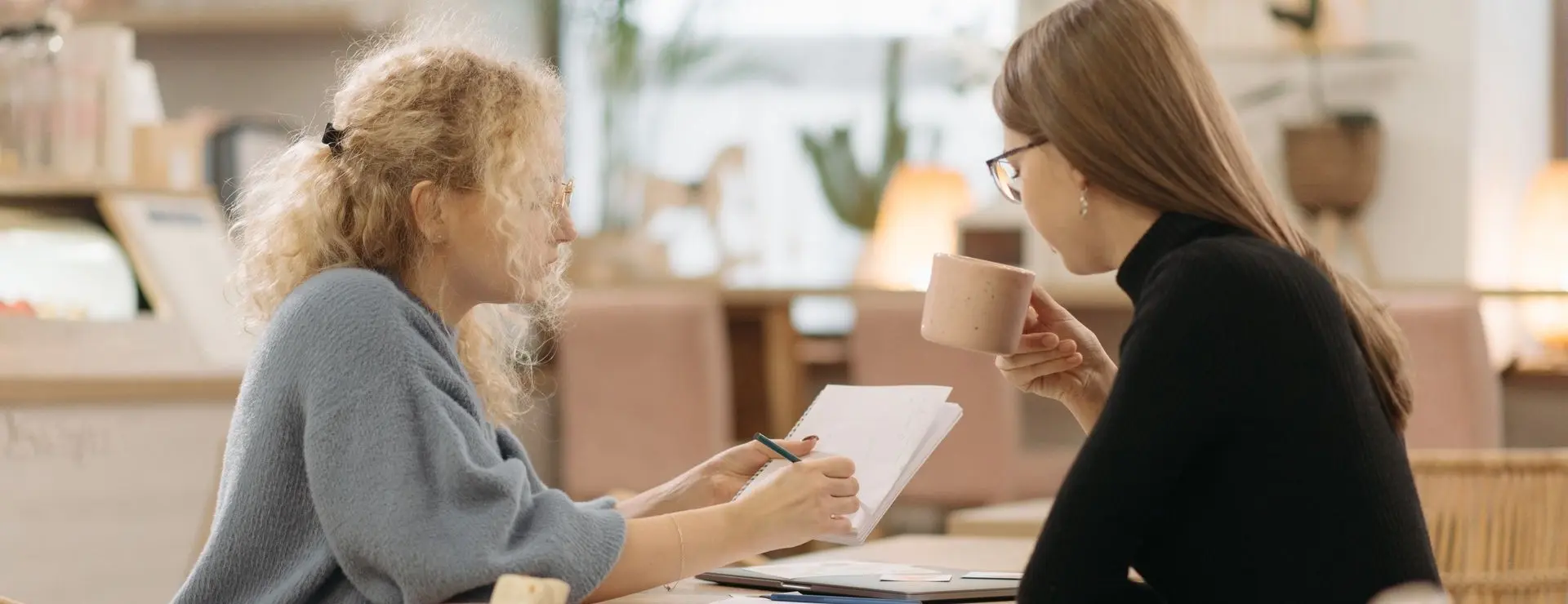 Two people sit at a table indoors, reading and sharing a takeaway cup while reviewing papers together.