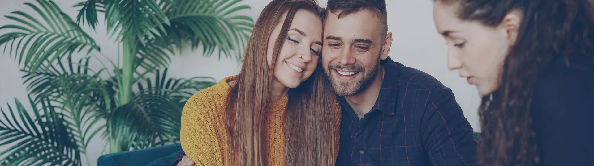 A smiling couple cuddles closely on a couch indoors, with large green palm leaves in the background; two other blurred people appear in the right foreground.