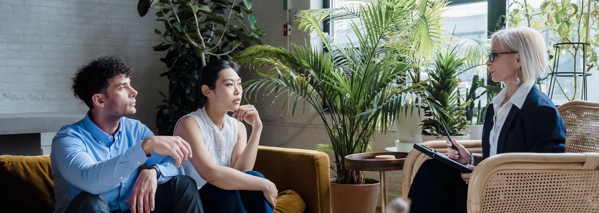 A meeting in a modern office lounge with three people seated near tall green plants; two clients sit talking while a woman in a blazer holds a tablet and listens.