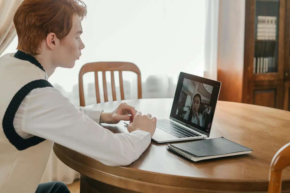 A man sitting at a wooden table using a laptop for a video call, seen in profile with another person displayed on the screen.